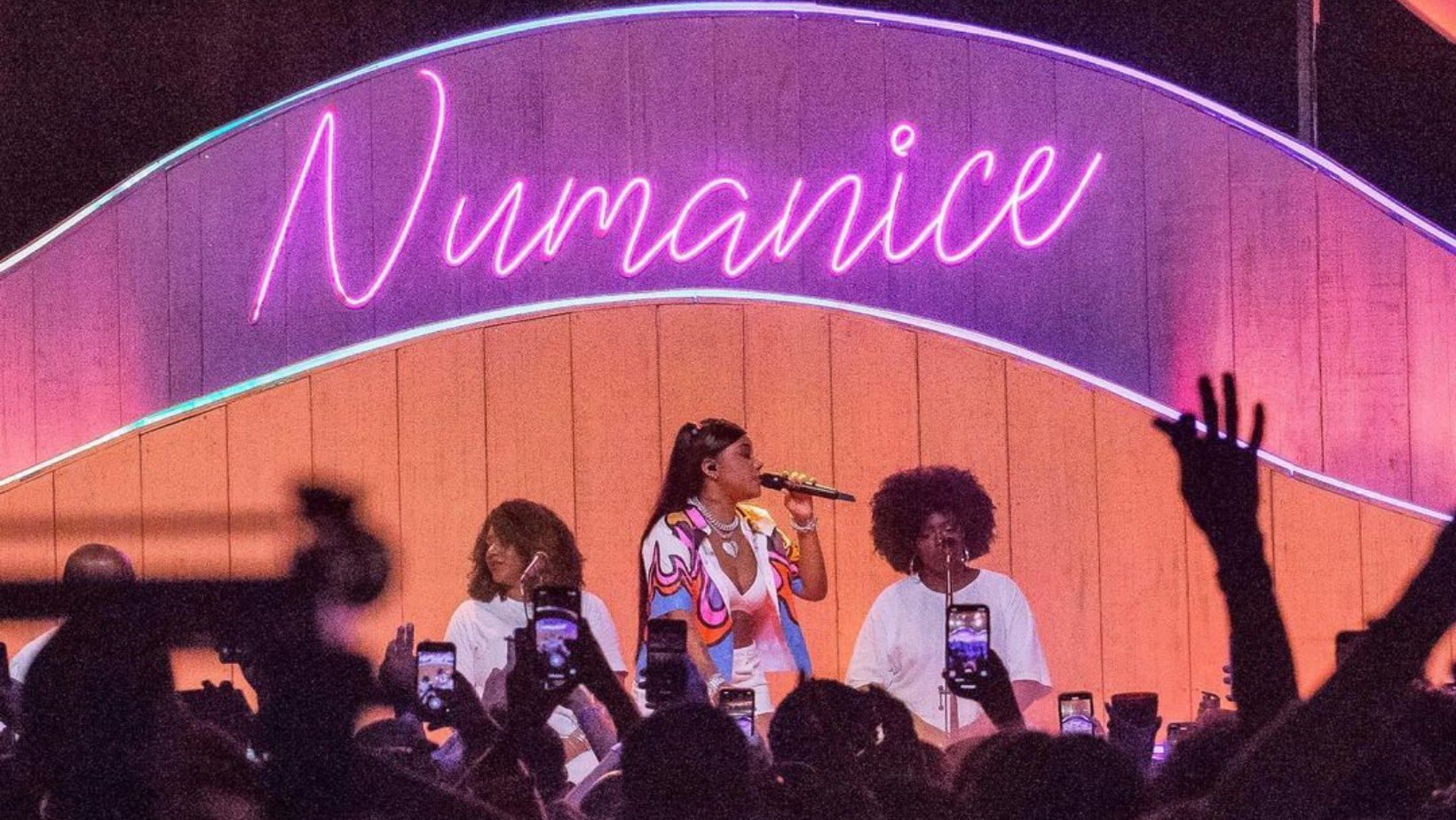 Imagem de Ludmilla segurando um microfone com um cenário de palco iluminado atrás dela